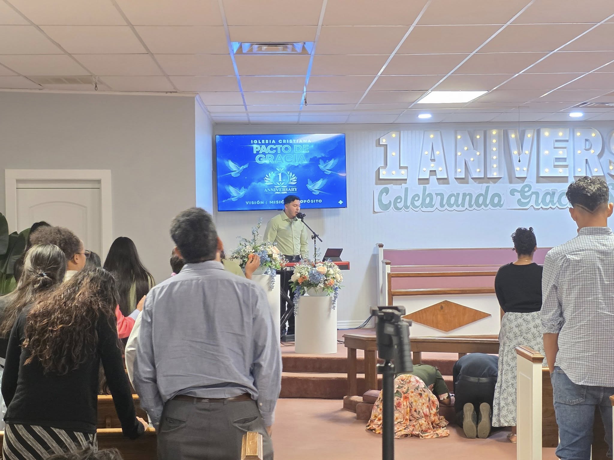 Celebración del primer aniversario de la Iglesia Cristiana Pacto de Gracia en Charlotte, con pastores y congregación reunidos en el púlpito decorado con flores y pantallas.
