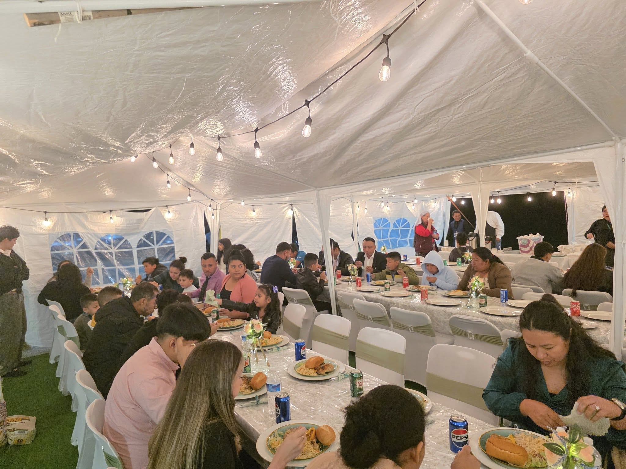 Congregación de la Iglesia Cristiana Pacto de Gracia en Charlotte compartiendo alimentos y bebidas en mesas largas bajo carpa decorada con luces, durante la celebración del primer aniversario.