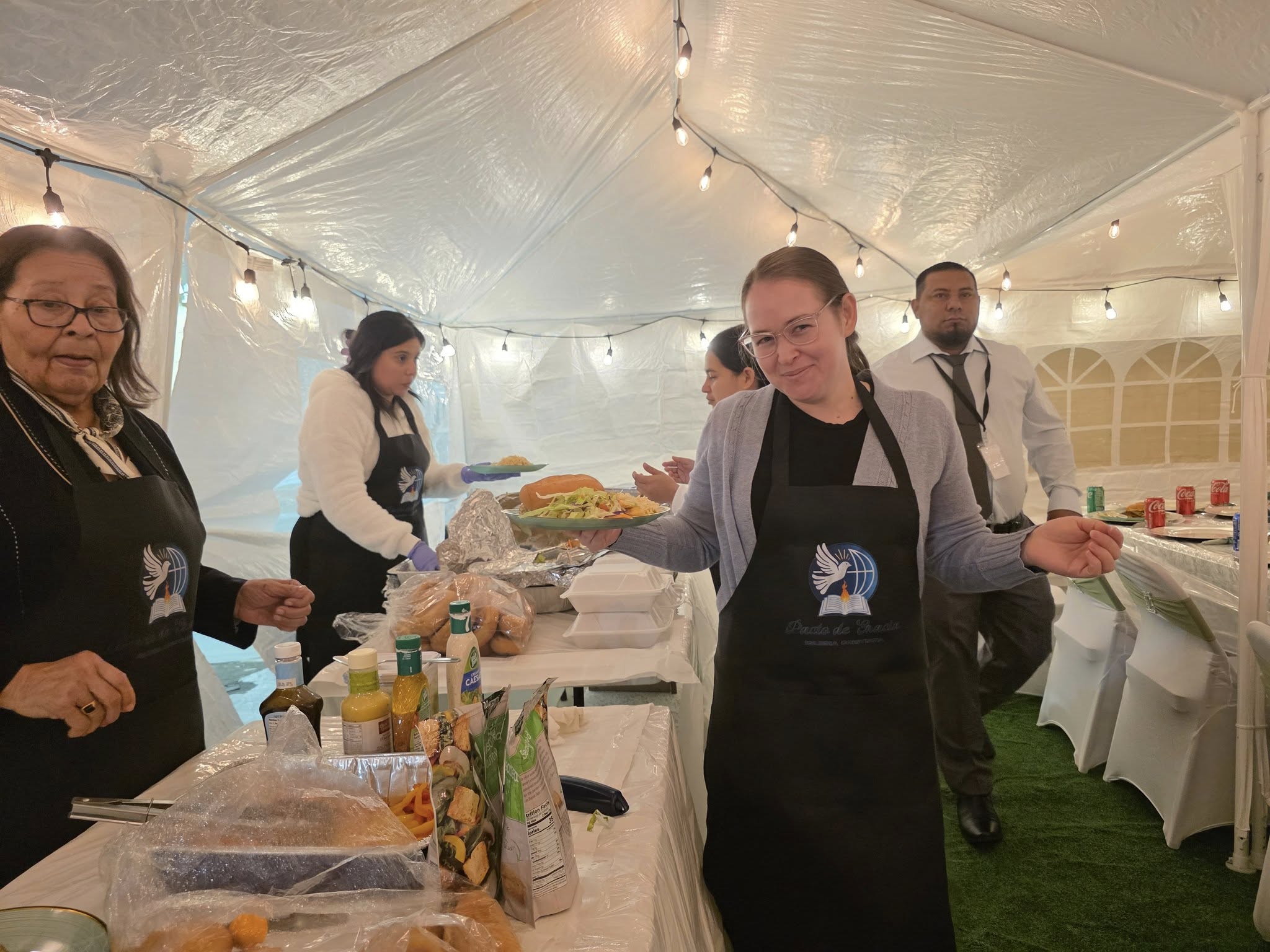 Mujeres de la Iglesia Cristiana Pacto de Gracia en Charlotte sirviendo alimentos y bebidas durante la celebración del primer aniversario, en un ambiente festivo bajo carpa decorada con luces.