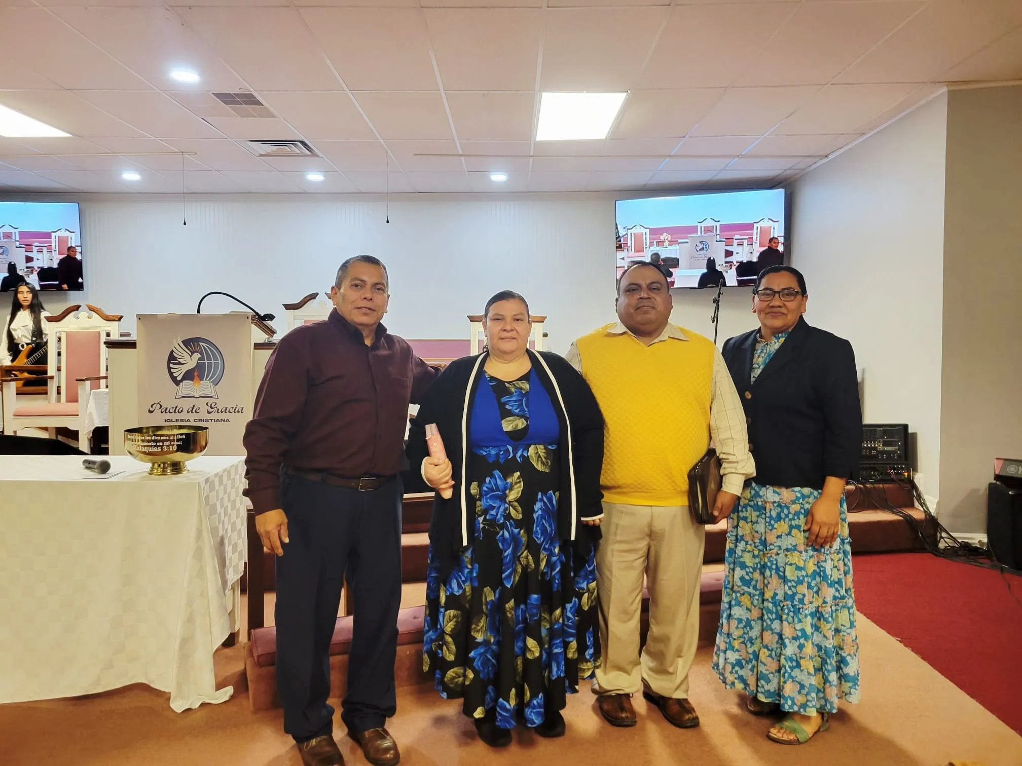 Líderes y miembros de la Iglesia Cristiana Pacto de Gracia en Charlotte participando en un servicio especial, vestidos formalmente frente al púlpito y pantallas con mensajes congregacionales.