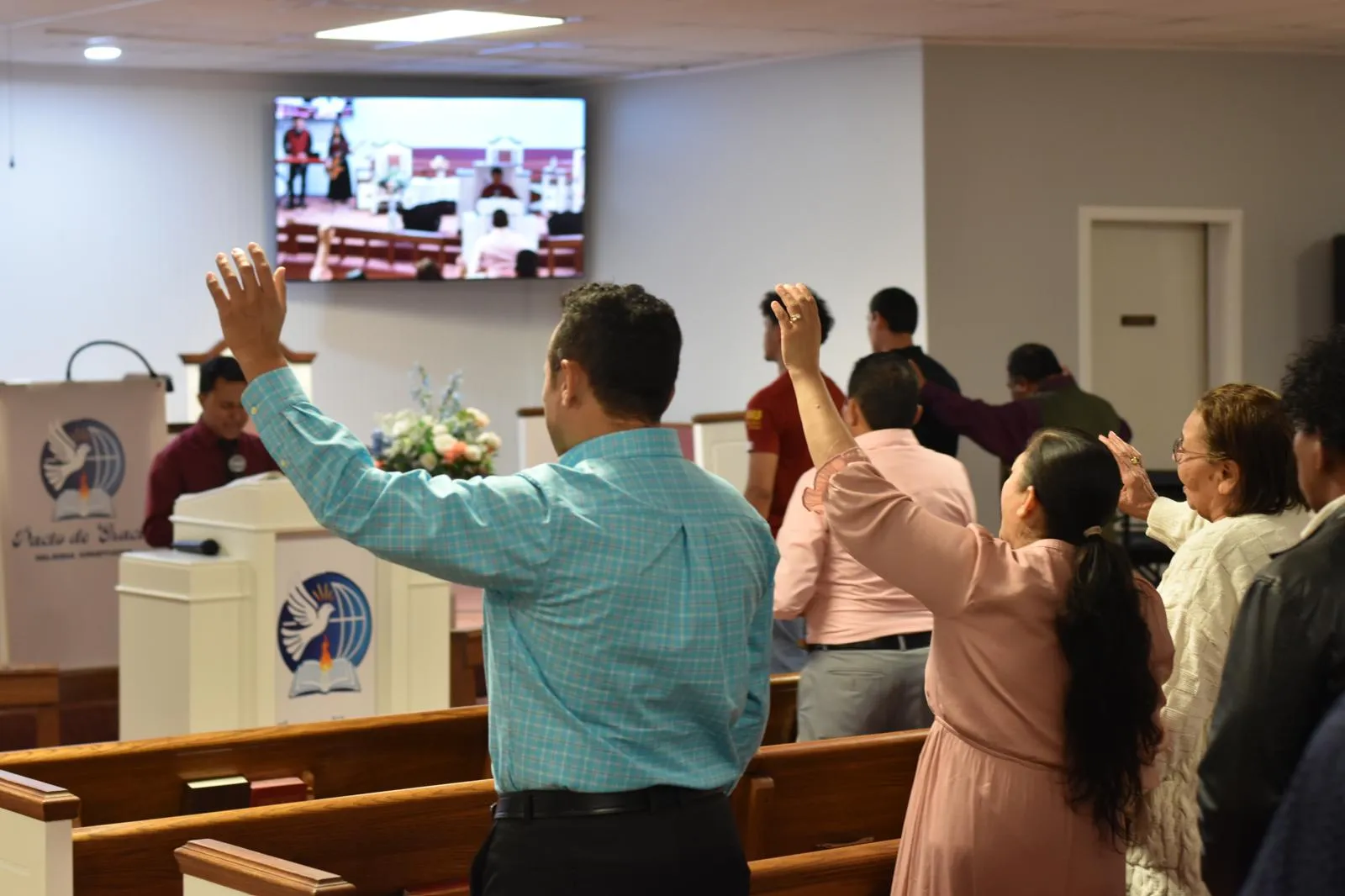 Congregación de la Iglesia Cristiana Pacto de Gracia en Charlotte participando en un servicio especial de adoración, con músicos al frente y el logo con globo y cruz en el púlpito.