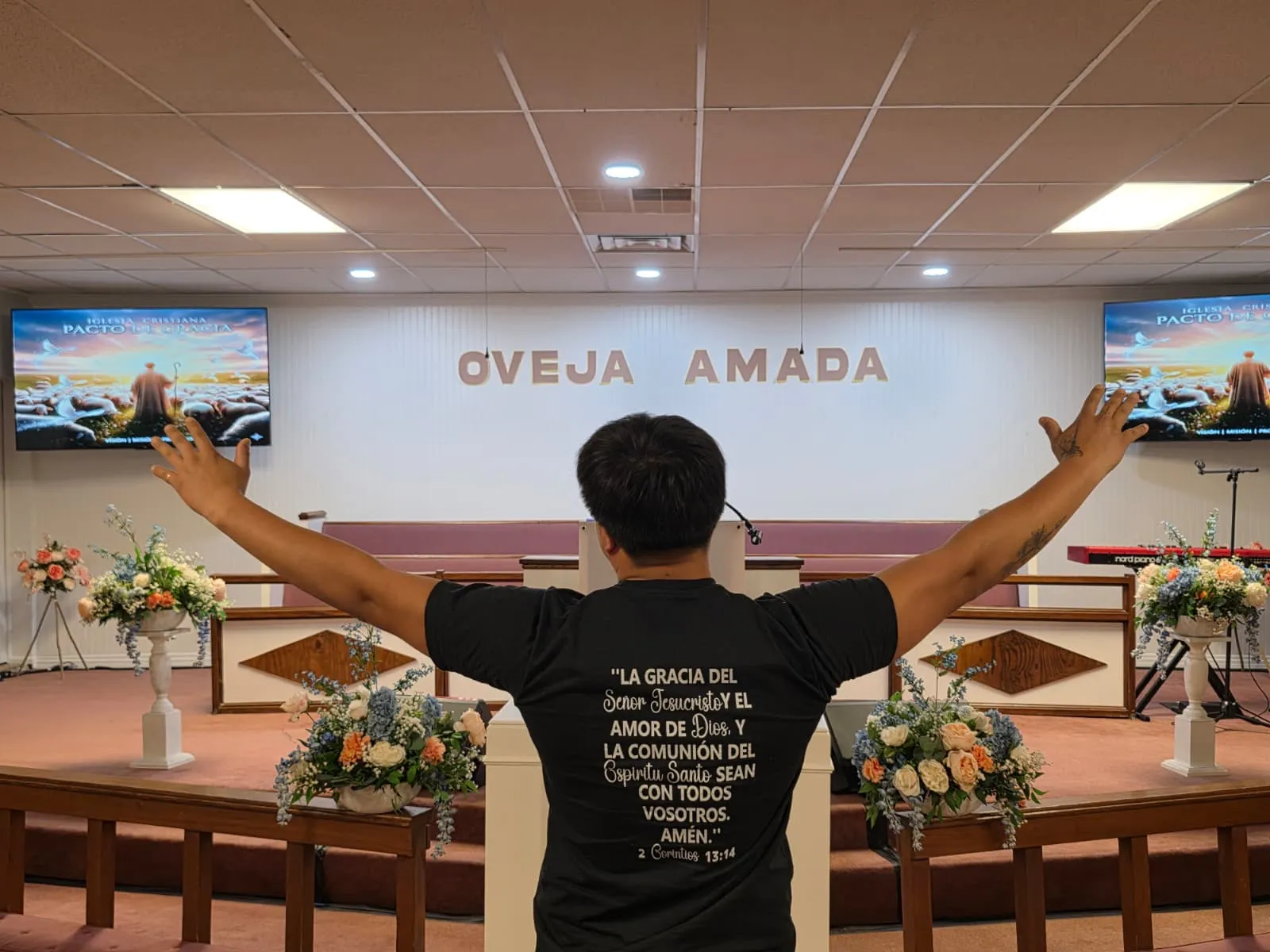 Miembro de la Iglesia Cristiana Pacto de Gracia en Charlotte levantando sus brazos en adoración frente al altar con la frase “Gloria a Dios”, vistiendo una camiseta con mensaje cristiano en la parte posterior.