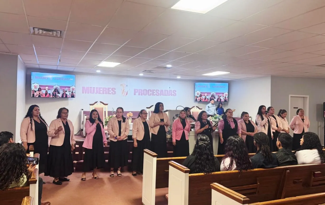 Grupo de mujeres de la Iglesia Cristiana Pacto de Gracia en Charlotte participando en el servicio especial Women’s Day, vestidas de manera coordinada con bufandas rosadas frente al púlpito.