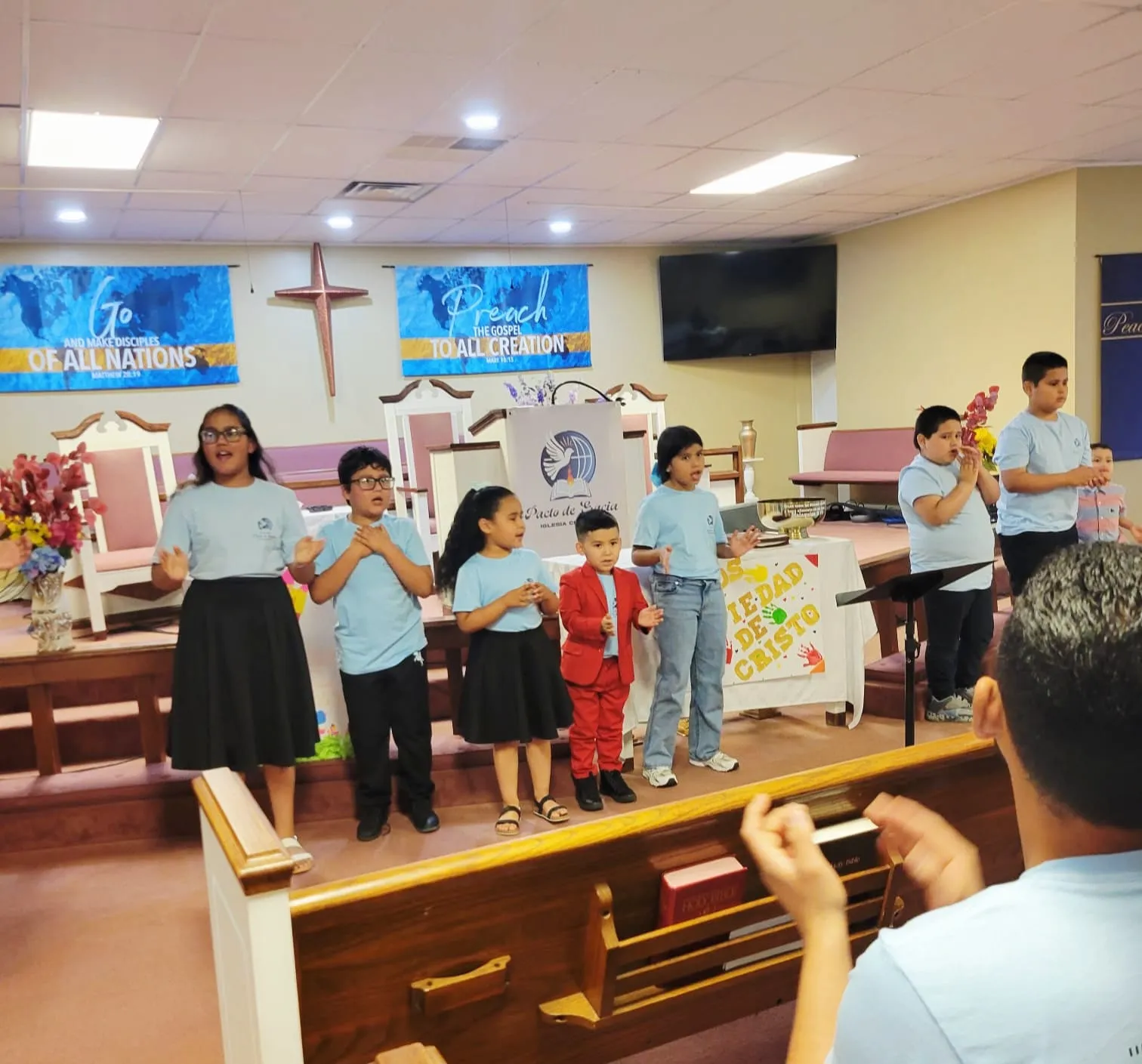 Niños de la Iglesia Cristiana Pacto de Gracia en Charlotte participando en una presentación congregacional, acompañados por adultos y con decoraciones alusivas a la fe y la comunidad.
