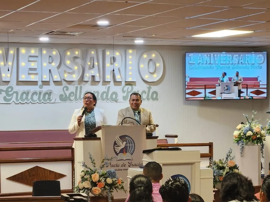 Pastores de la Iglesia Cristiana Pacto de Gracia en Charlotte durante el culto del primer aniversario, en el púlpito con flores y congregación presente.
